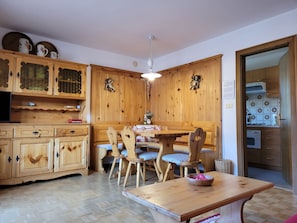 Een keuken met houten kastjes, een eettafel met stoelen, een plafondlamp en uitzicht op de aangrenzende kamer.