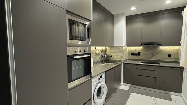 Une cuisine moderne avec des meubles de rangement de couleur sombre, un four encastré, un four à micro-ondes et un lave-linge.