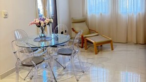 Un salon moderne avec une table à manger en verre, des chaises transparentes, un canapé beige et une grande fenêtre avec des rideaux.