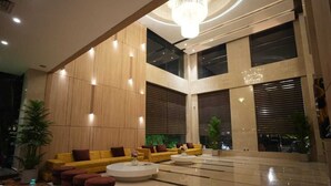 Un hall d’entrée spacieux au design moderne, avec un espace de détente central agrémenté de canapés jaunes et de poufs rouges, le tout entouré d’un sol poli et de grandes fenêtres.