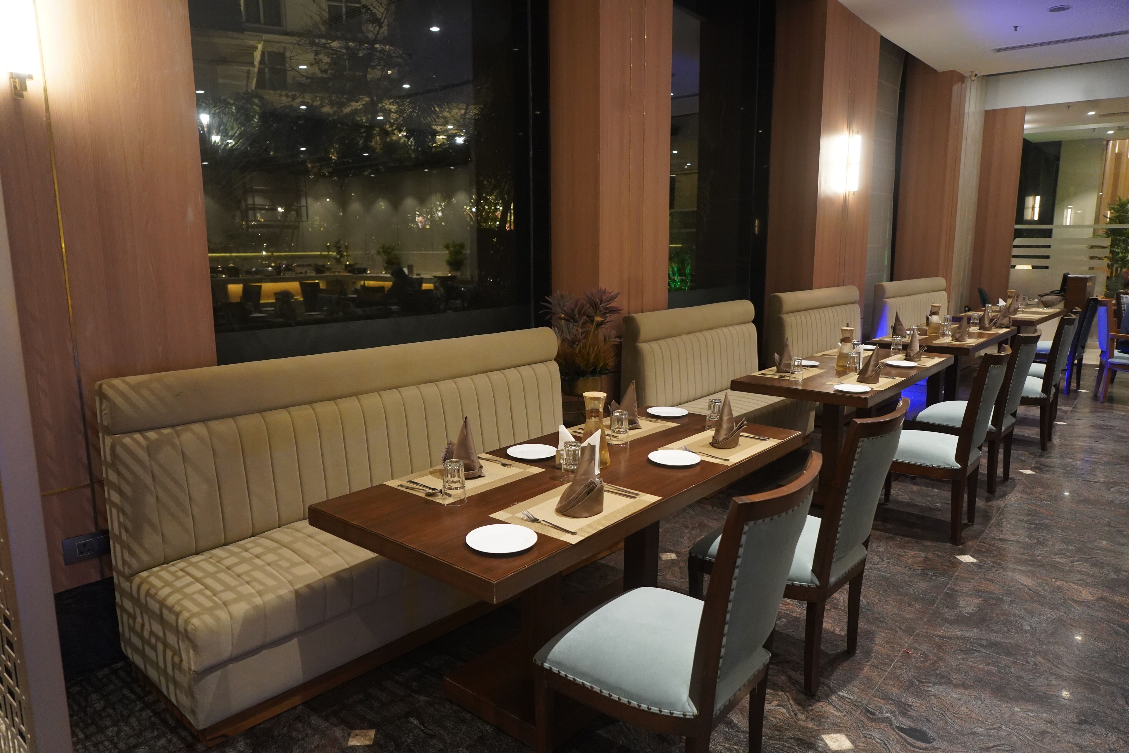 Ein Restaurant mit einem langen Holztisch, gedeckten Plätzen, bequemen Sitzgelegenheiten und Blick auf die beleuchtete Stadt bei Nacht.