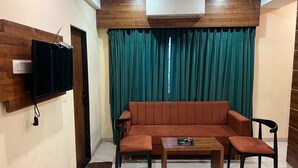 Ein Wohnzimmer mit einer Couch, zwei Sesseln, einem Couchtisch, einem flachbildschirmfernseher an der Wand und einem großen Fenster mit türkisfarbenen Vorhängen.