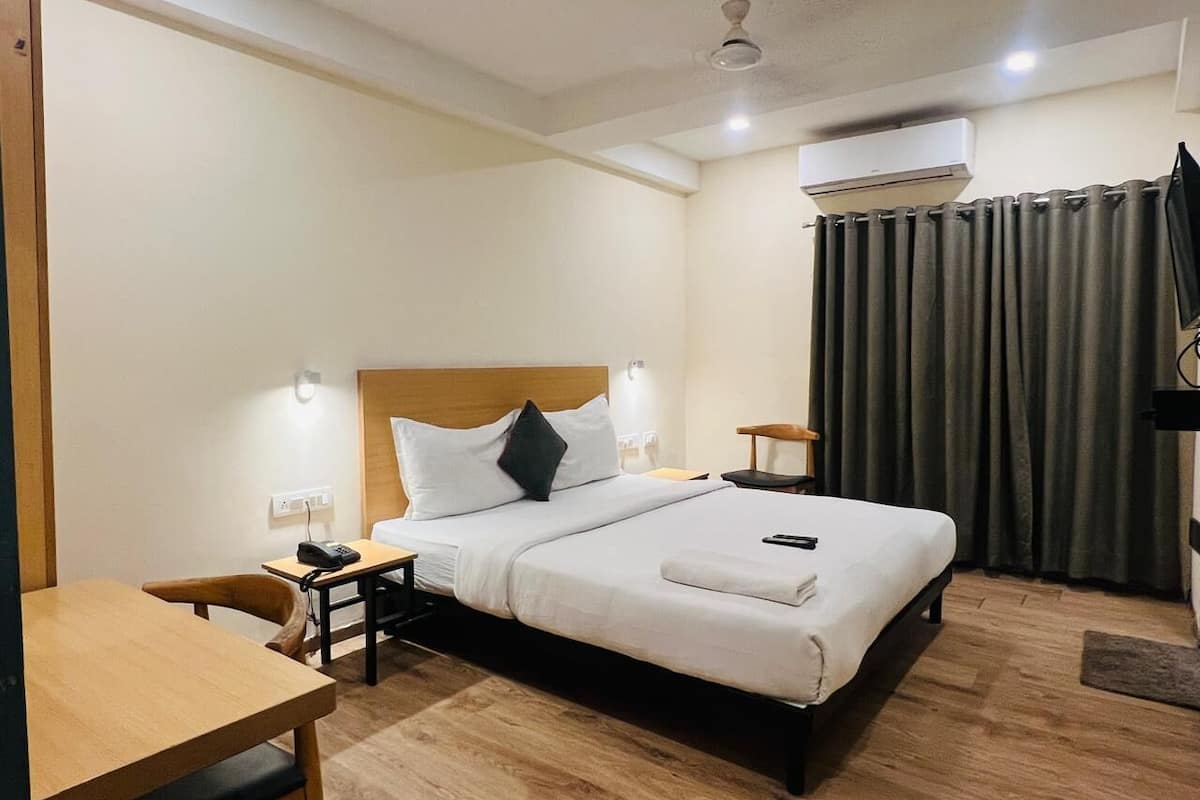 A hotel room with a bed, bedside table, chair, desk, and an air conditioning unit.