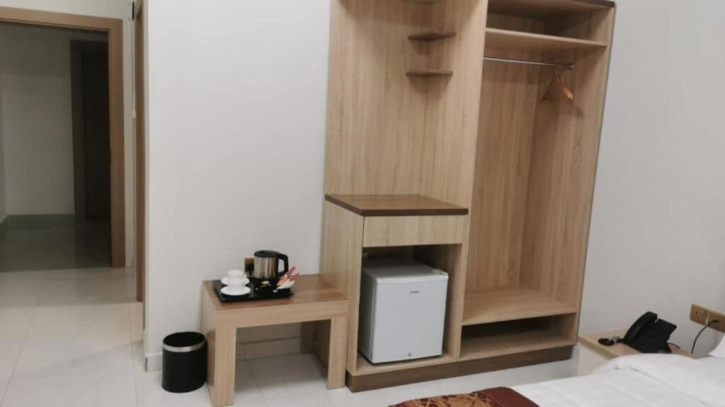 A hotel room with a wooden wardrobe, a small table with a coffee maker, a mini fridge, a trash bin, and a bed.