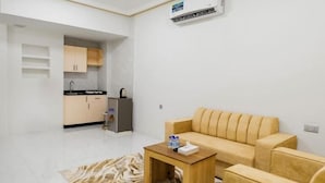 Ein modernes Wohnzimmer mit einem beigefarbenen Sofa, einem Holztisch und einem Teppich mit Muster. Im Hintergrund ist eine Küchenzeile mit Schränken und einem Kühlschrank zu sehen.