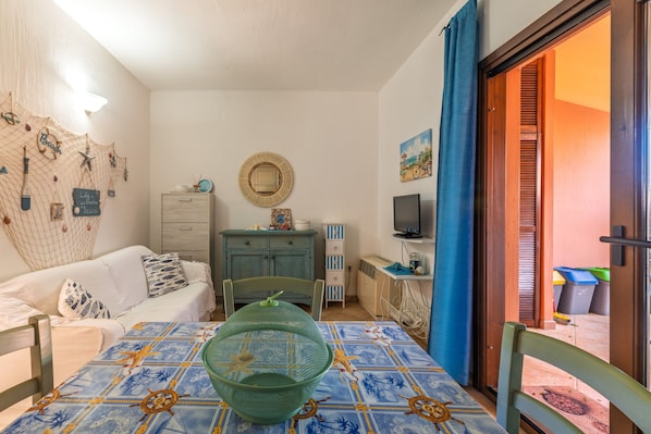 Une pièce comprenant un lit, une table recouverte d’une nappe à motifs, une chaise verte, un meuble en bois, un téléviseur et une porte donnant sur un espace extérieur.