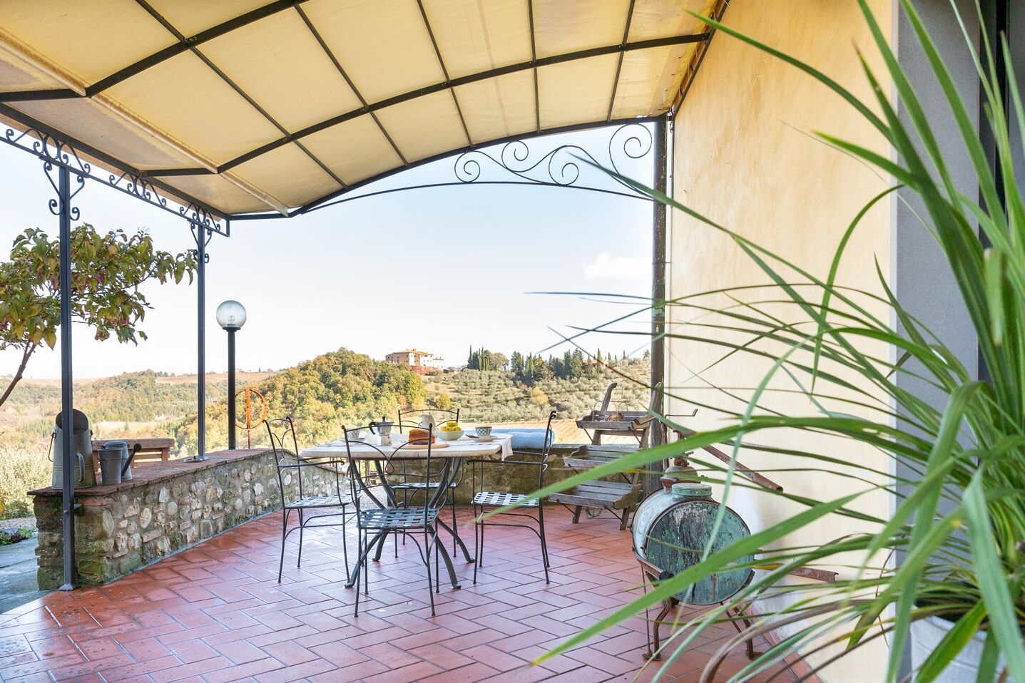 A covered outdoor patio with stone walls, metal chairs, a table, and a view of a hillside.