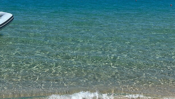 Une plage à l’eau turquoise et limpide, un rivage sablonneux et plusieurs bateaux visibles au loin.