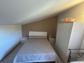 Una habitación pequeña con cama individual, techo inclinado, mesita de noche con lámpara y armario.