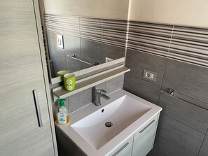Un baño con un lavabo blanco, un espejo y una estantería con dos tazas verdes.