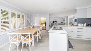 A modern kitchen with white cabinetry, a central island with a sink, and a dining area with a wooden table and white chairs. - Serenity - A place for relaxing (Corlette)