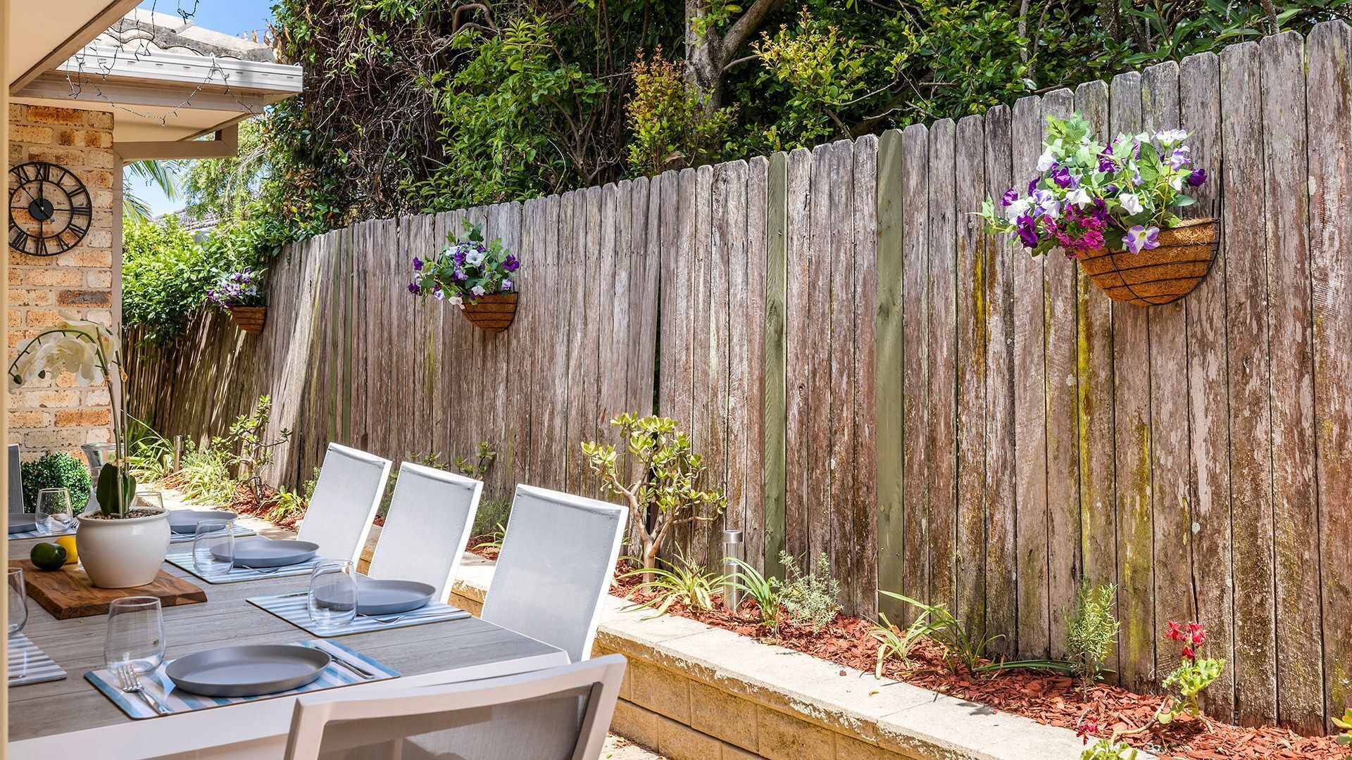 An outdoor dining area with a wooden fence, hanging flower baskets, and a clock on the wall.