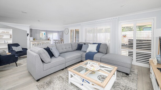 A modern living room with a grey sectional sofa, a glass coffee table, and a kitchen in the background. - Serenity - A place for relaxing (Corlette)