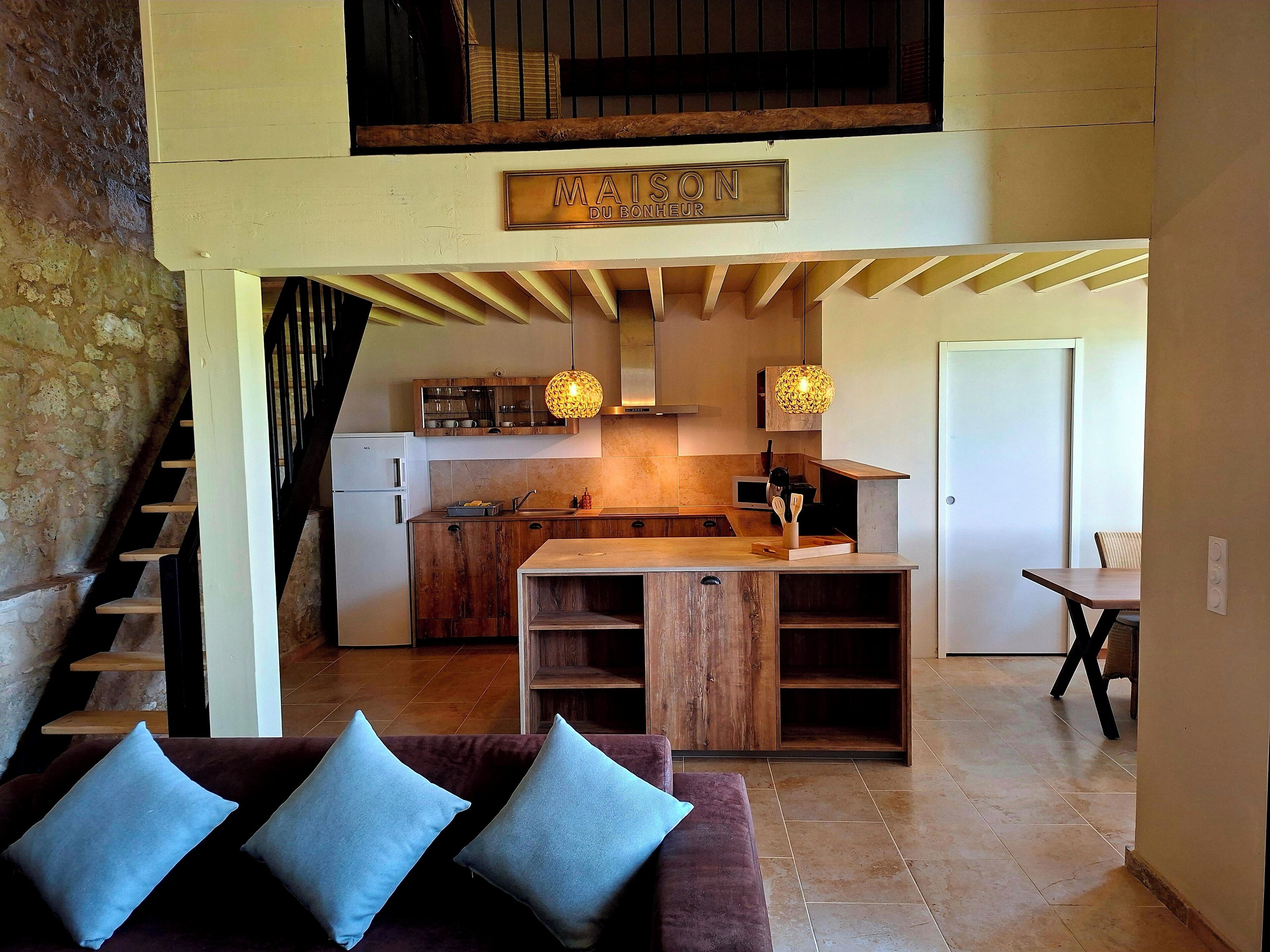 Une cuisine moderne avec un îlot central en bois, un coin repas avec une table et des chaises, un escalier et un salon avec un canapé et des coussins.