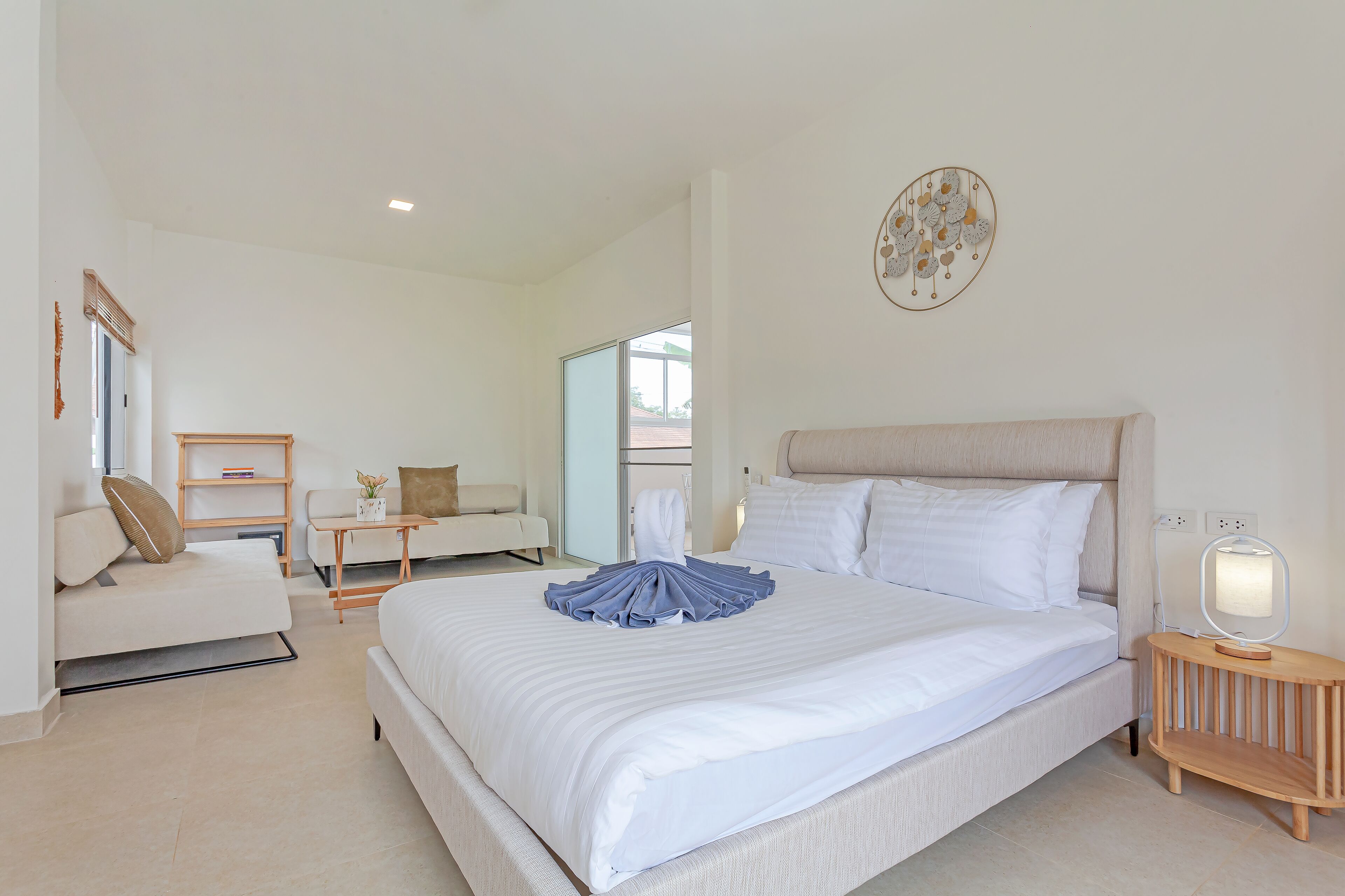 A modern hotel room with a large bed, a bedside table with a lamp, a small seating area with a sofa and a table, and a large window providing a view of the outside.
