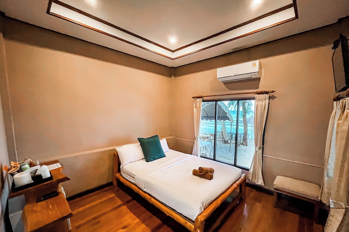 A bedroom with a wooden bed, a nightstand, an air conditioning unit, and a view of the beach through the window.