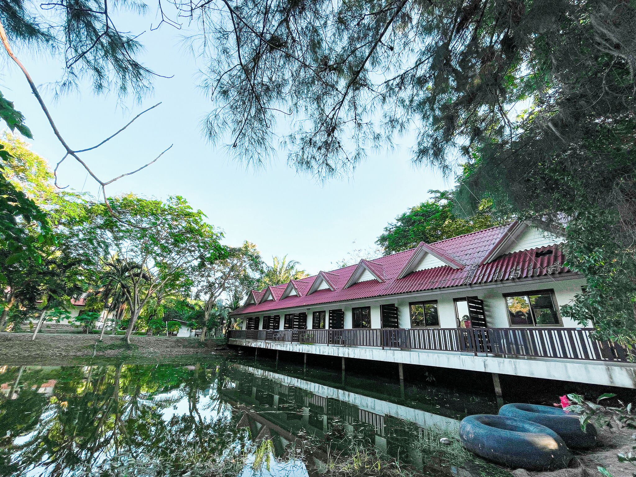 A building with a red roof and white walls, situated next to a calm body of water, surrounded by trees.