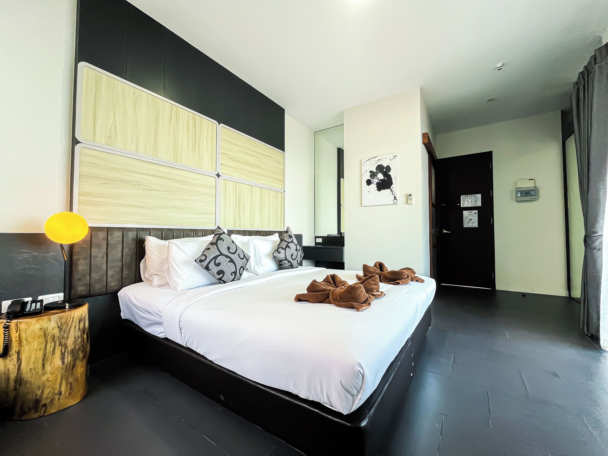 A modern hotel room with a large bed, a bedside lamp, a wooden side table, and a black door.