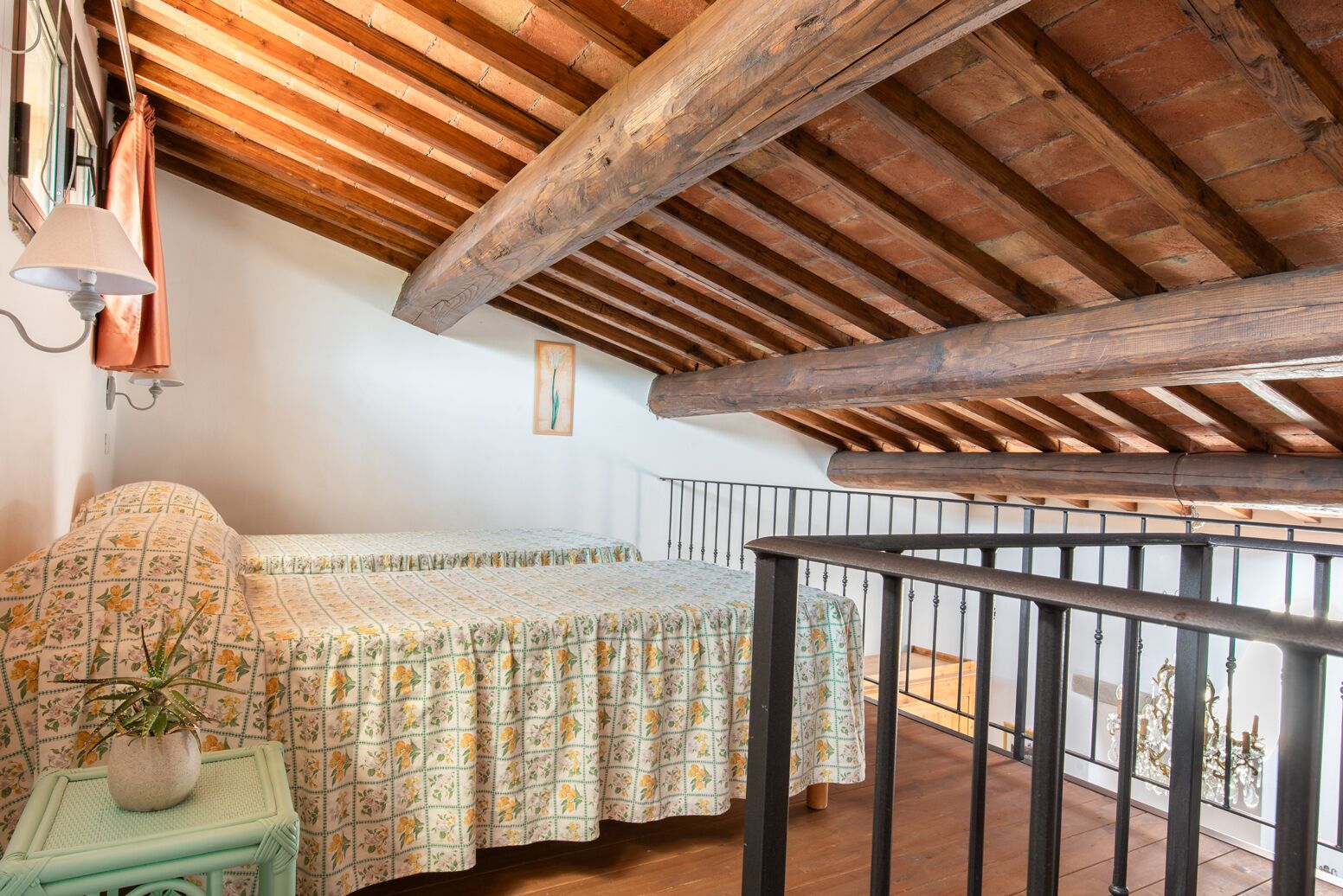 Ein Schlafzimmer mit Holzboden, einem Bett mit floraler Bettwäsche, einem kleinen Tisch mit einer Pflanze und einem Balkongeländer.