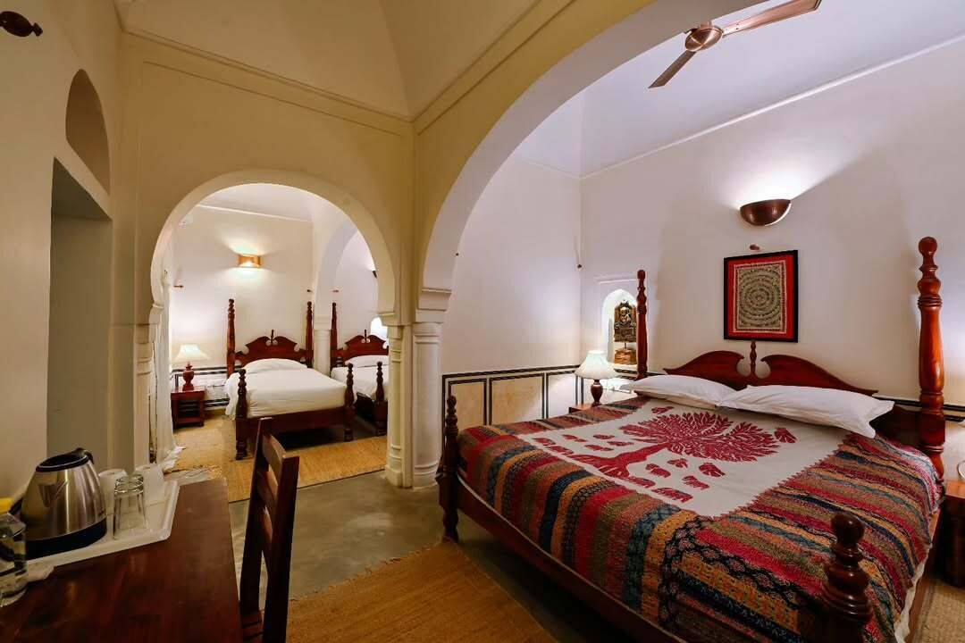 Une chambre d’hôtel avec un grand lit, un bureau en bois et une chaise. Il y a un ventilateur de plafond et un tableau encadré au mur.