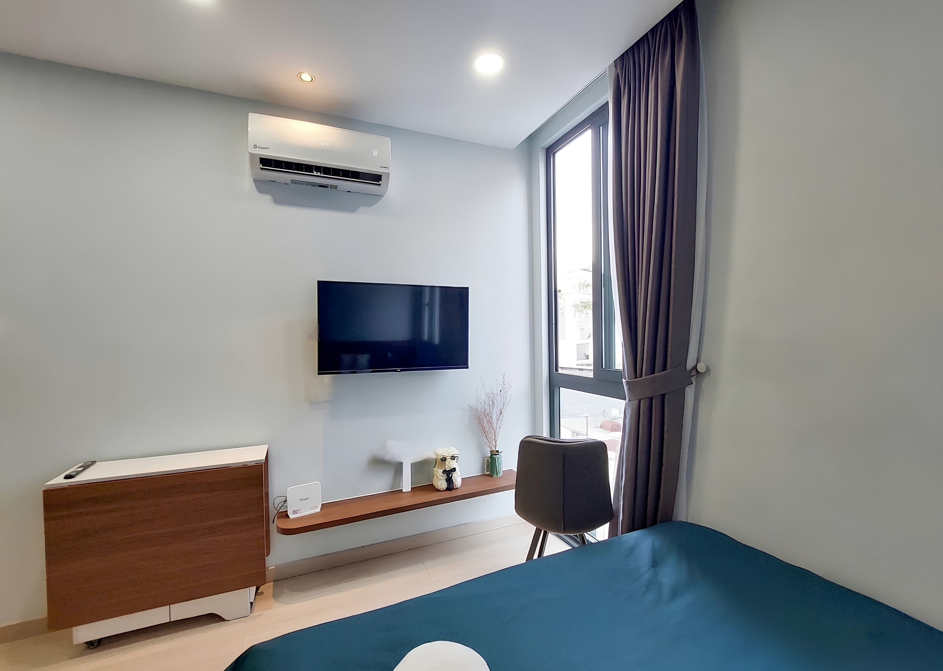 Un dormitorio moderno con una televisión de pantalla plana, un equipo de aire acondicionado, un aparador de madera, una mesita de noche con lámpara y una ventana con cortinas.