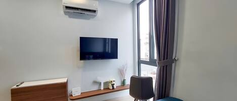 Un dormitorio moderno con una televisión de pantalla plana, un equipo de aire acondicionado, un aparador de madera, una mesita de noche con lámpara y una ventana con cortinas.