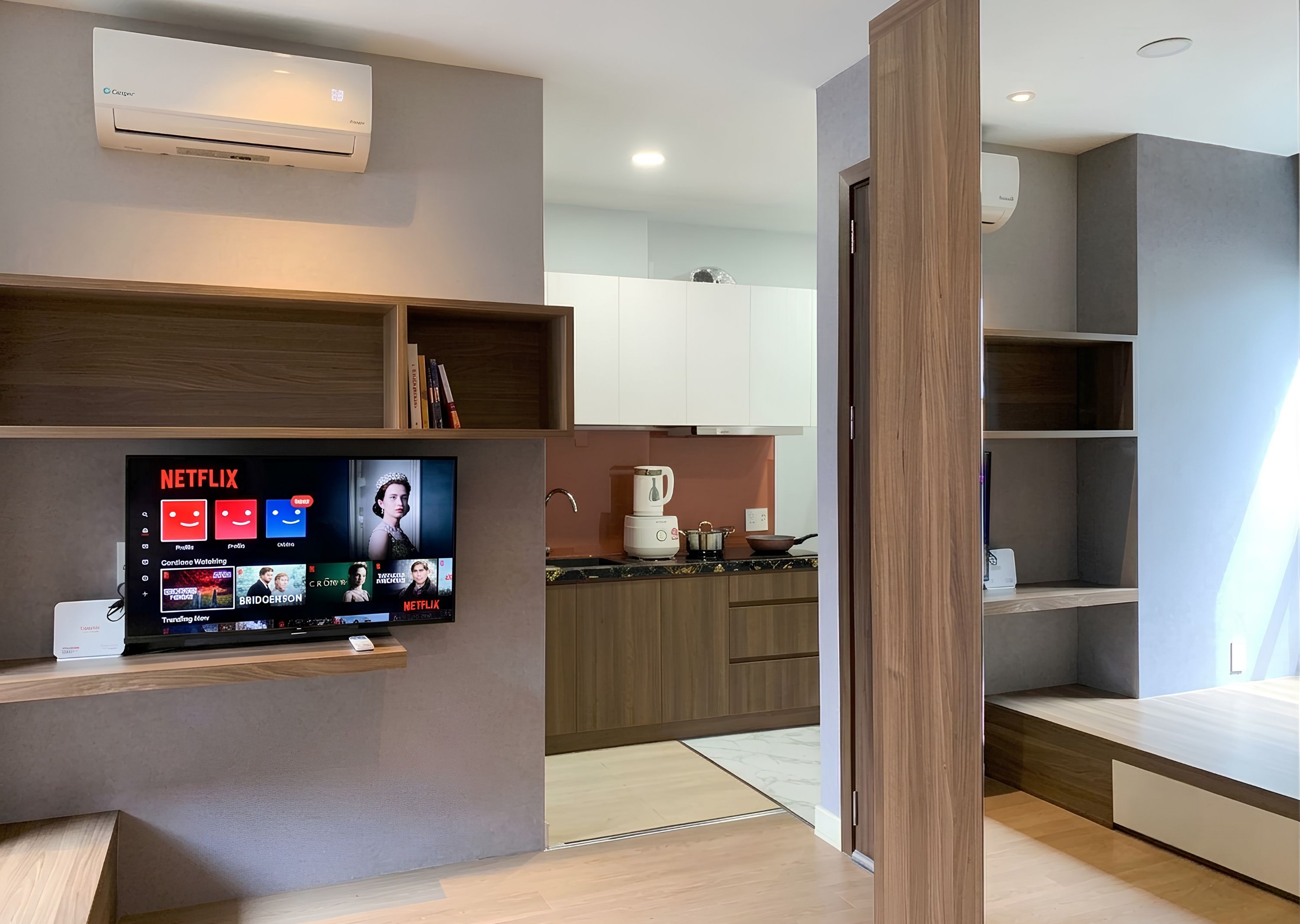 A modern living room with a flat-screen TV displaying Netflix content, a built-in air conditioner, and a minimalist design with wooden shelves and a kitchen area in the background.
