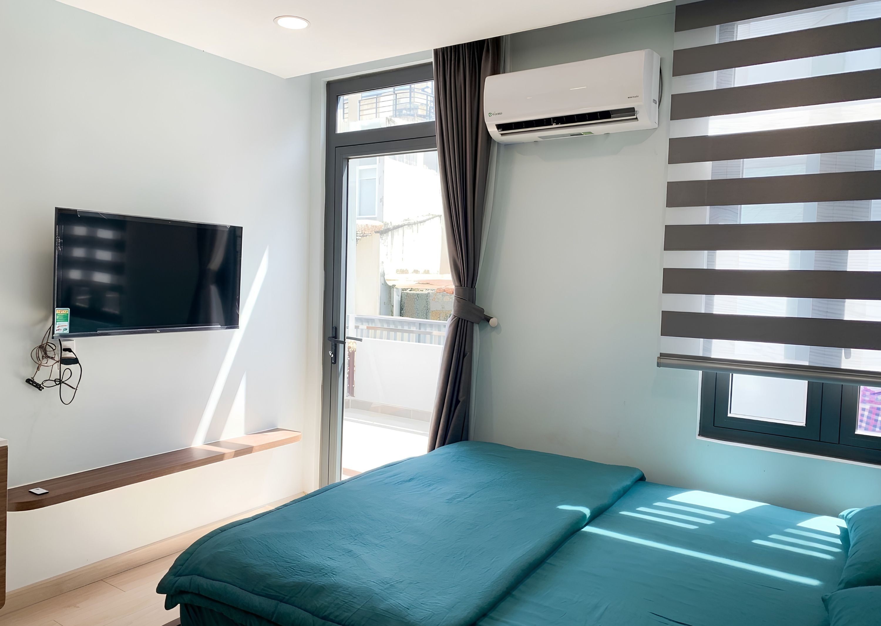 A bedroom with a bed, a mounted TV, an air conditioning unit, and a window with blinds.