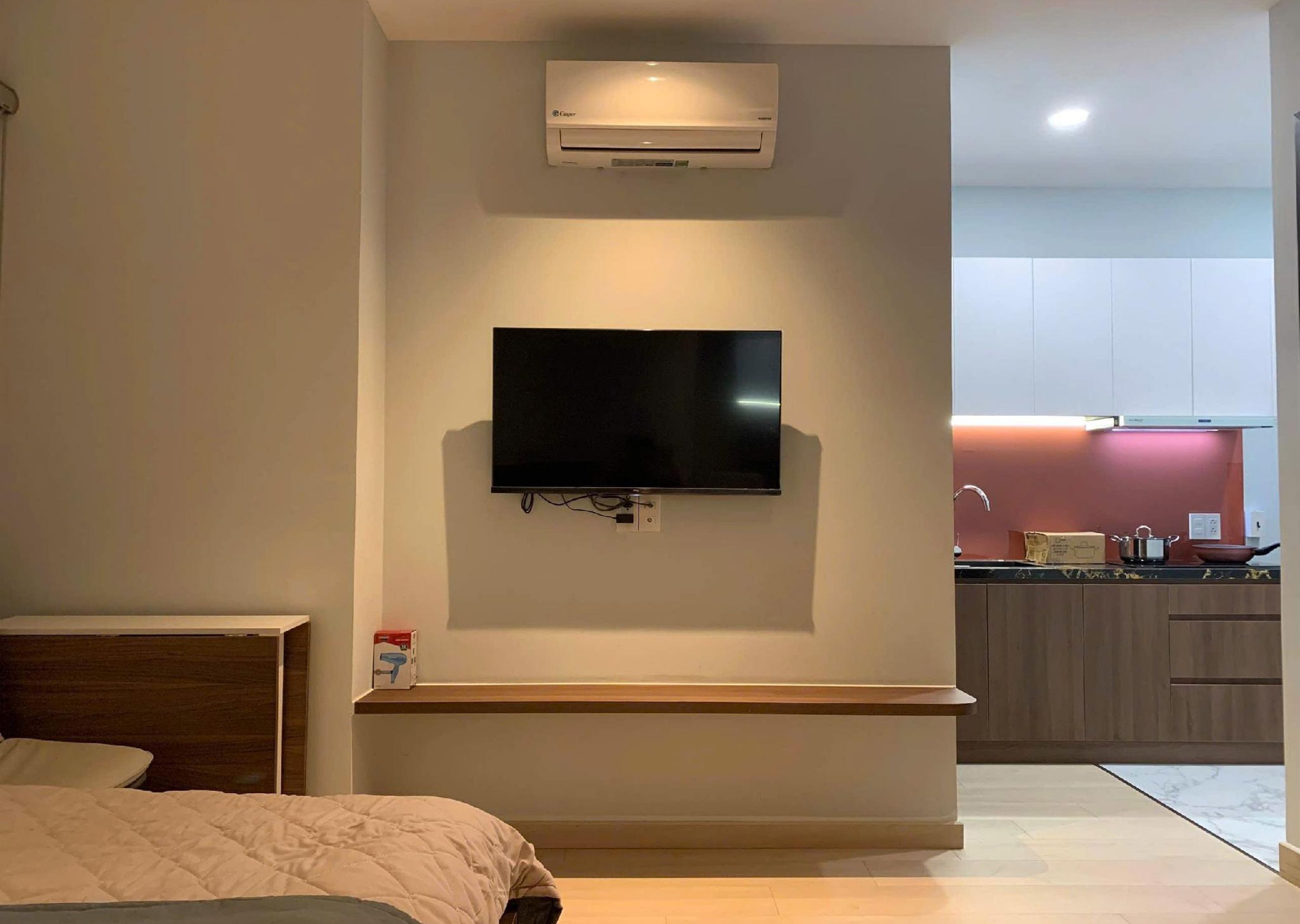 A modern hotel room with a flat-screen TV mounted on the wall, a bedside table, a bed, and a kitchenette area in the background.