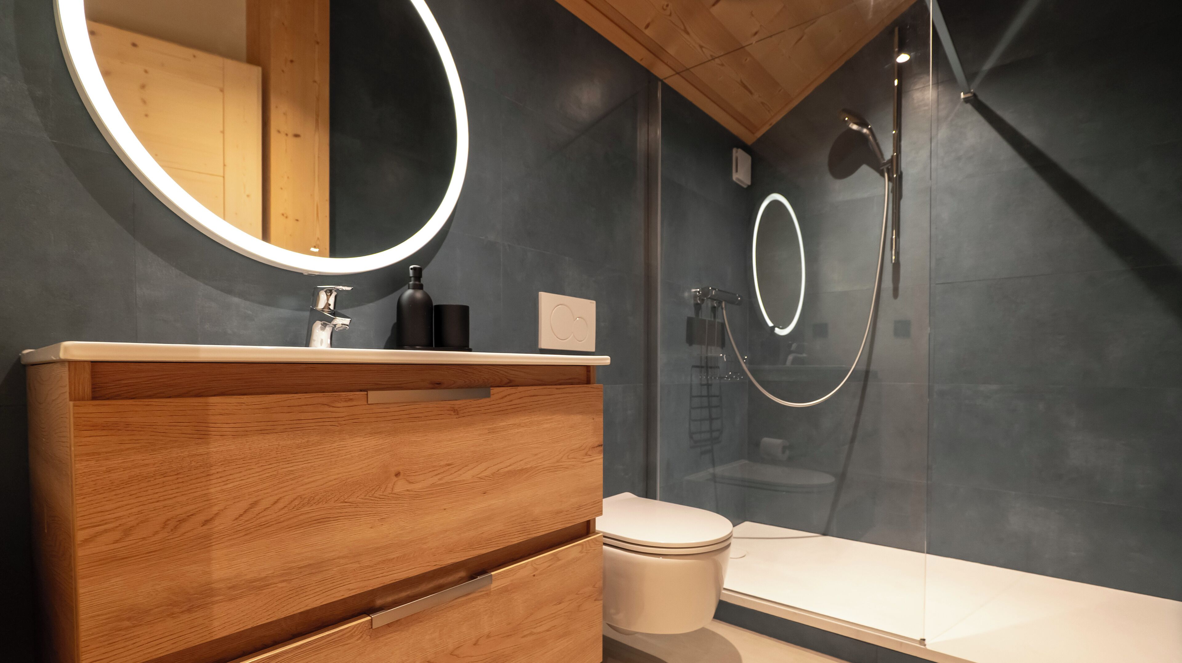 A modern bathroom with a wooden vanity, a round mirror, a toilet, and a glass-enclosed shower area.