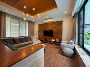 A modern living room with a wooden ceiling, a large flat-screen TV mounted on a wooden wall, a sofa, a coffee table, and a window with curtains. - Baan Wang Tan 1 Luxury Pool Villa, Chiang Mai (Chiang Mai)