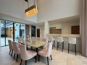 Un comedor moderno con una mesa de vidrio rodeada por seis sillas, una araña y una zona de cocina con taburetes de bar.