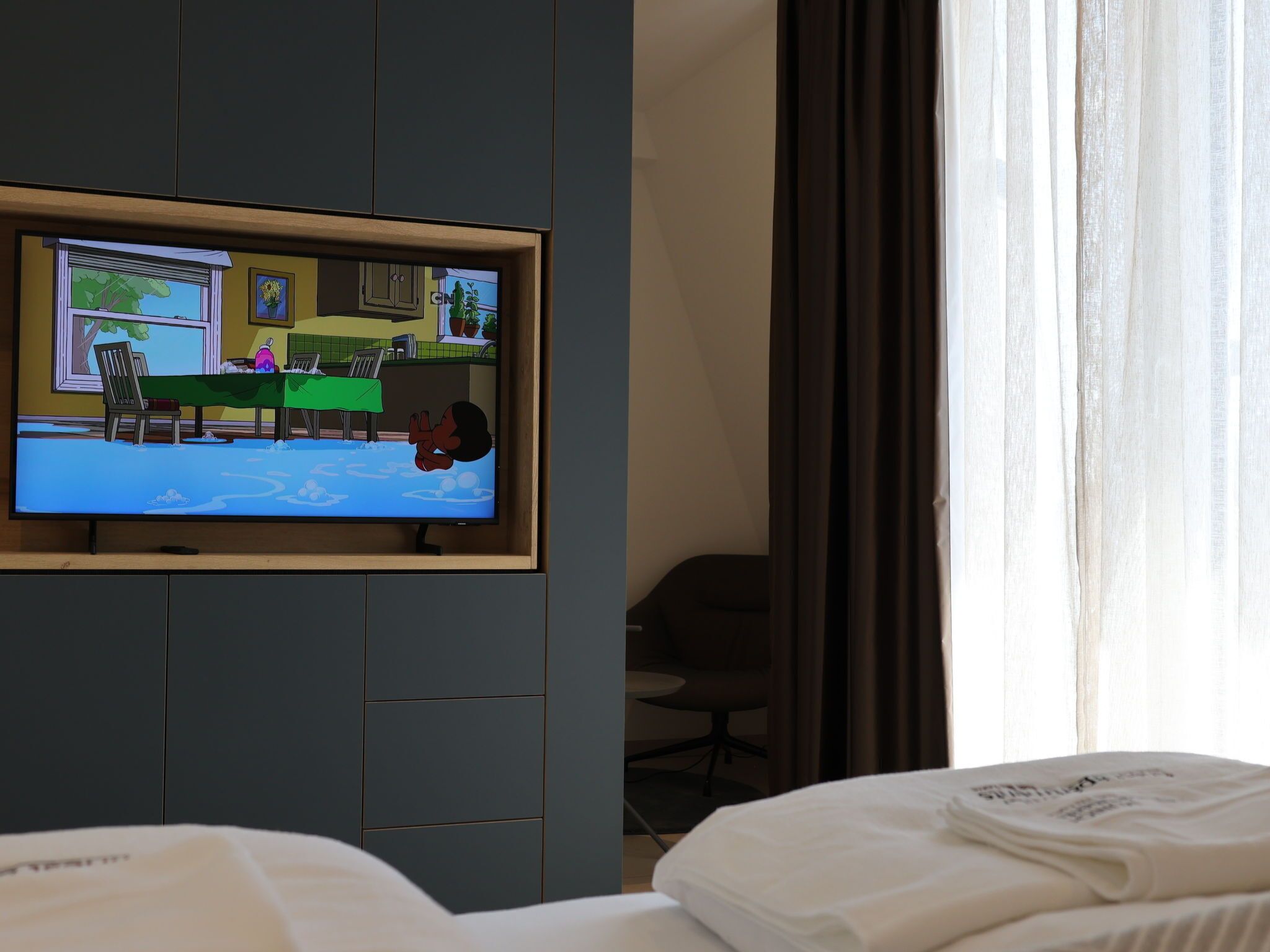 Een hotelkamer met een aan de muur bevestigde televisie, een bed met wit beddengoed, een bureau en een stoel.