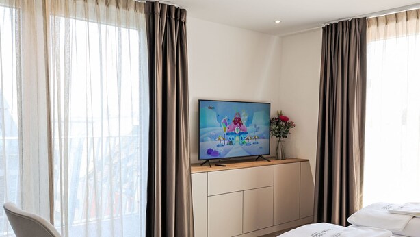 A hotel room with a TV on a wooden console, a white sofa, a small white table, and a large window with sheer curtains. - Munich Finest Apartments 20 by Interhome (Munich)