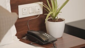 A neatly made bed with a white pillow, a bedside telephone, and a potted plant on a wooden nightstand. - Hotel Rajputana International Raipur (Raipur)