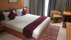 A hotel room with a large bed, a wooden headboard, a desk, a chair, and a view of the outside through curtains. - Hotel Rajputana International Raipur (Raipur)