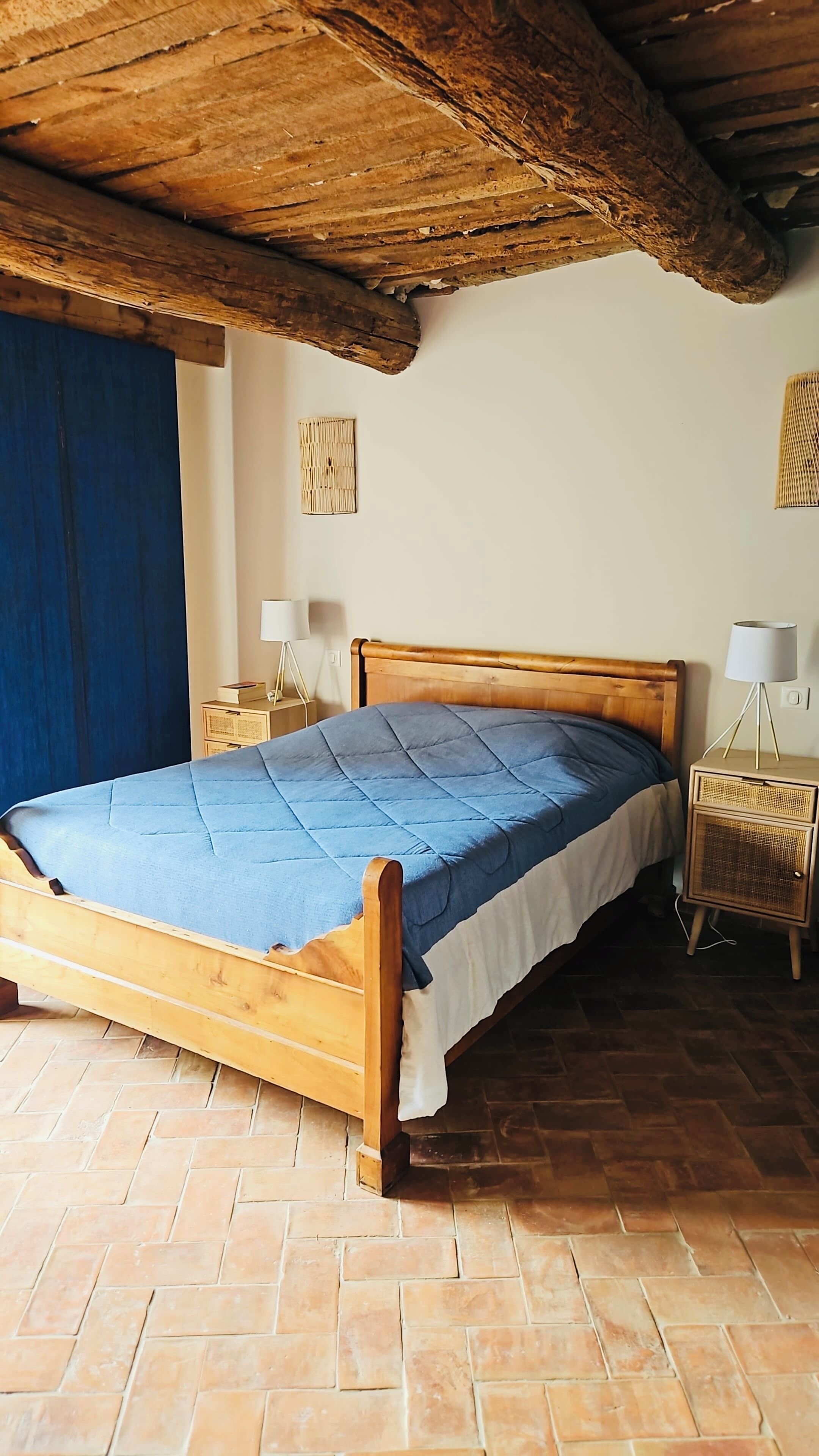 A wooden bed with a blue quilt, a bedside lamp, and a wicker nightstand in a room with a tiled floor and a wooden ceiling.