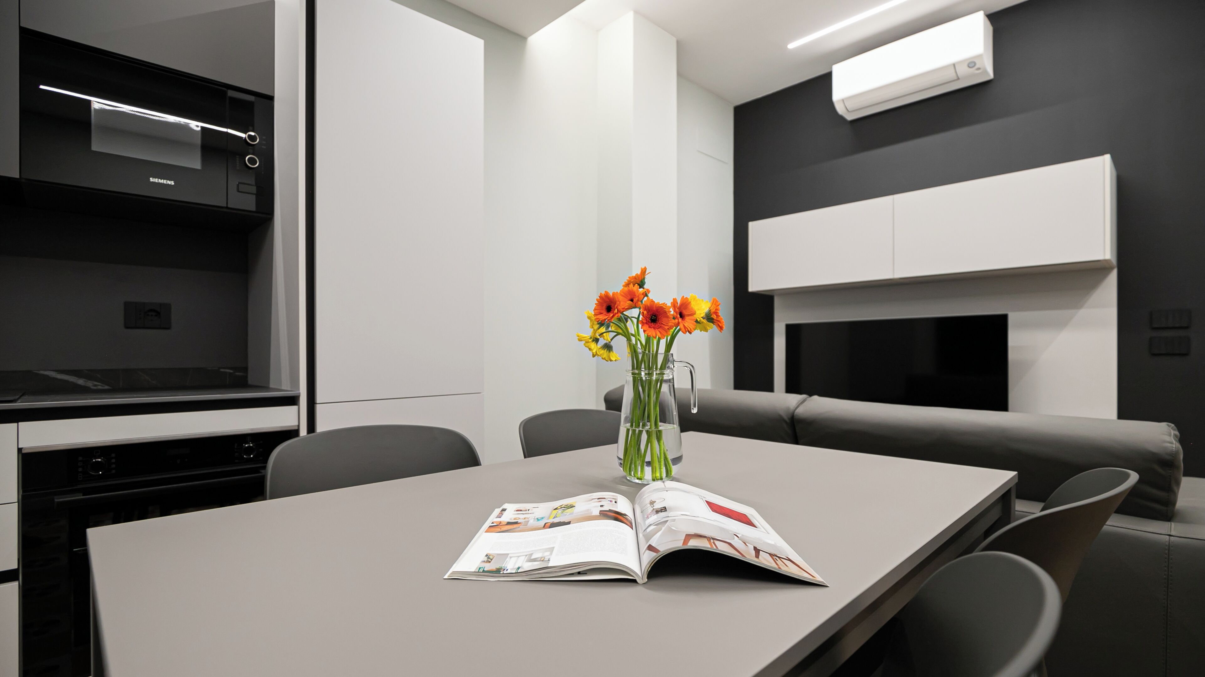 Una cocina moderna con un electrodoméstico Siemens, una mesa comedor gris con una revista y un florero, y un sofá gris.