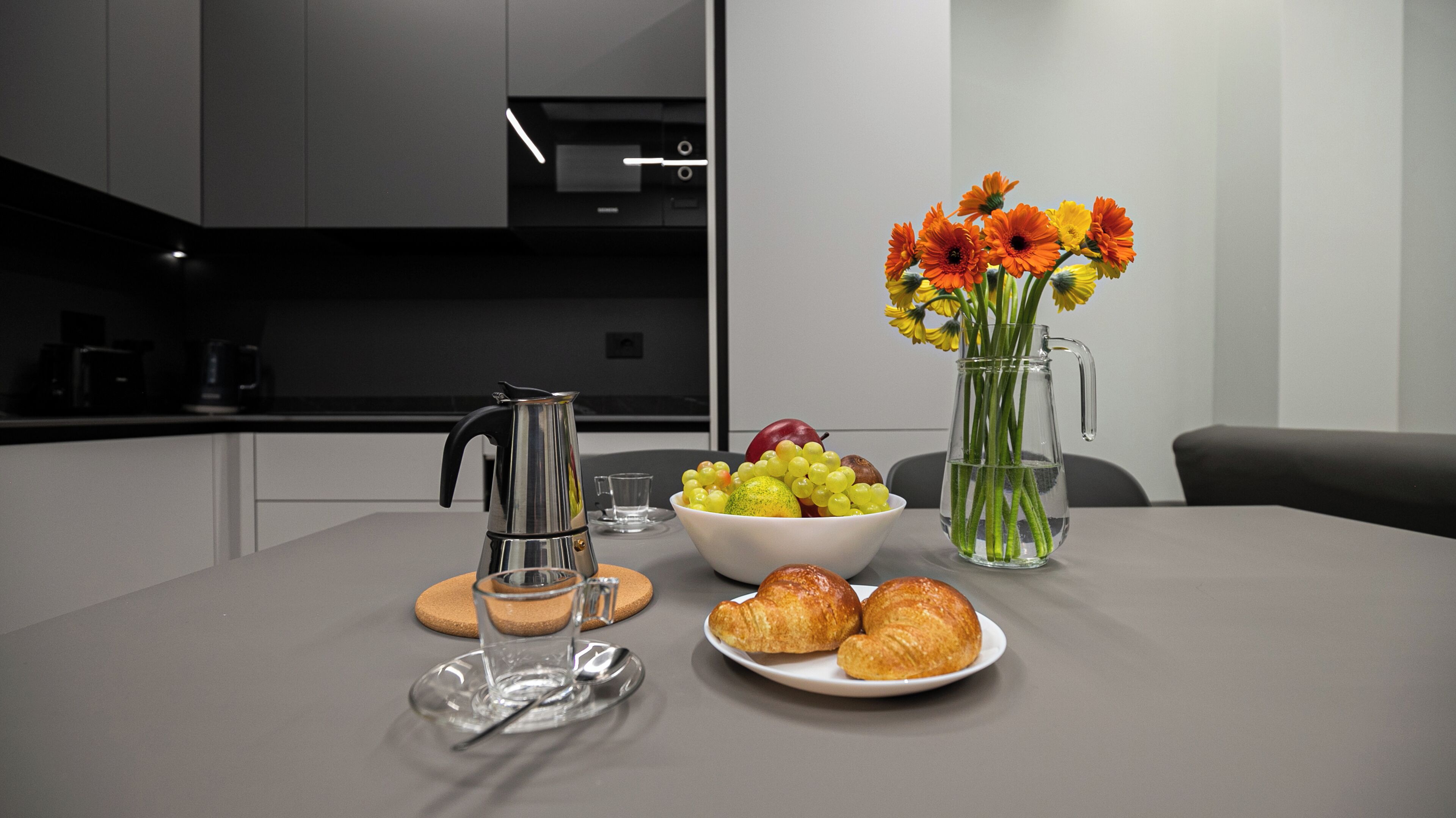 A modern kitchen with a coffee maker, a bowl of fruit, a vase with flowers, and a plate of croissants.