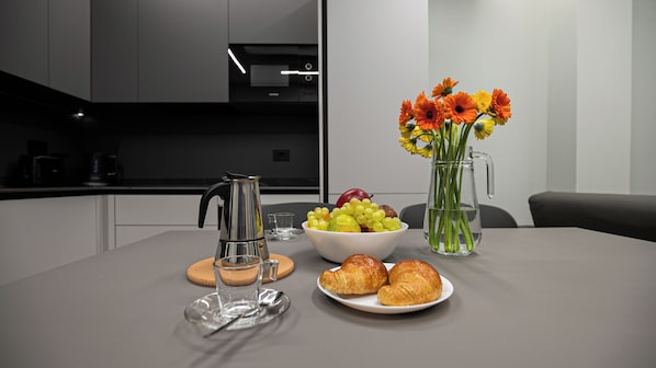 A modern kitchen with a coffee maker, a bowl of fruit, a vase with flowers, and a plate of croissants.