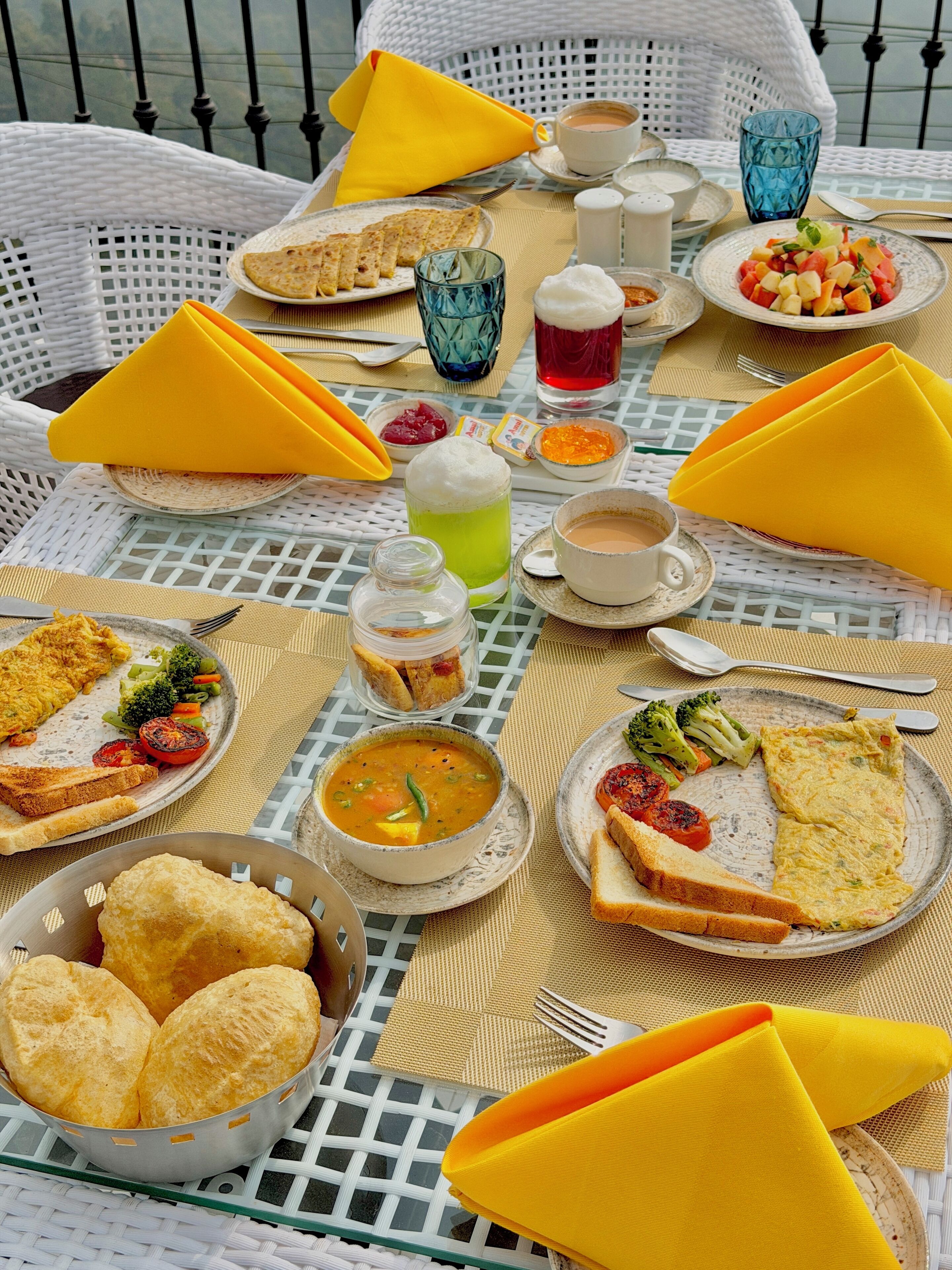 A table set with various dishes, including soup, toast, and a fruit salad, on a white wicker table.