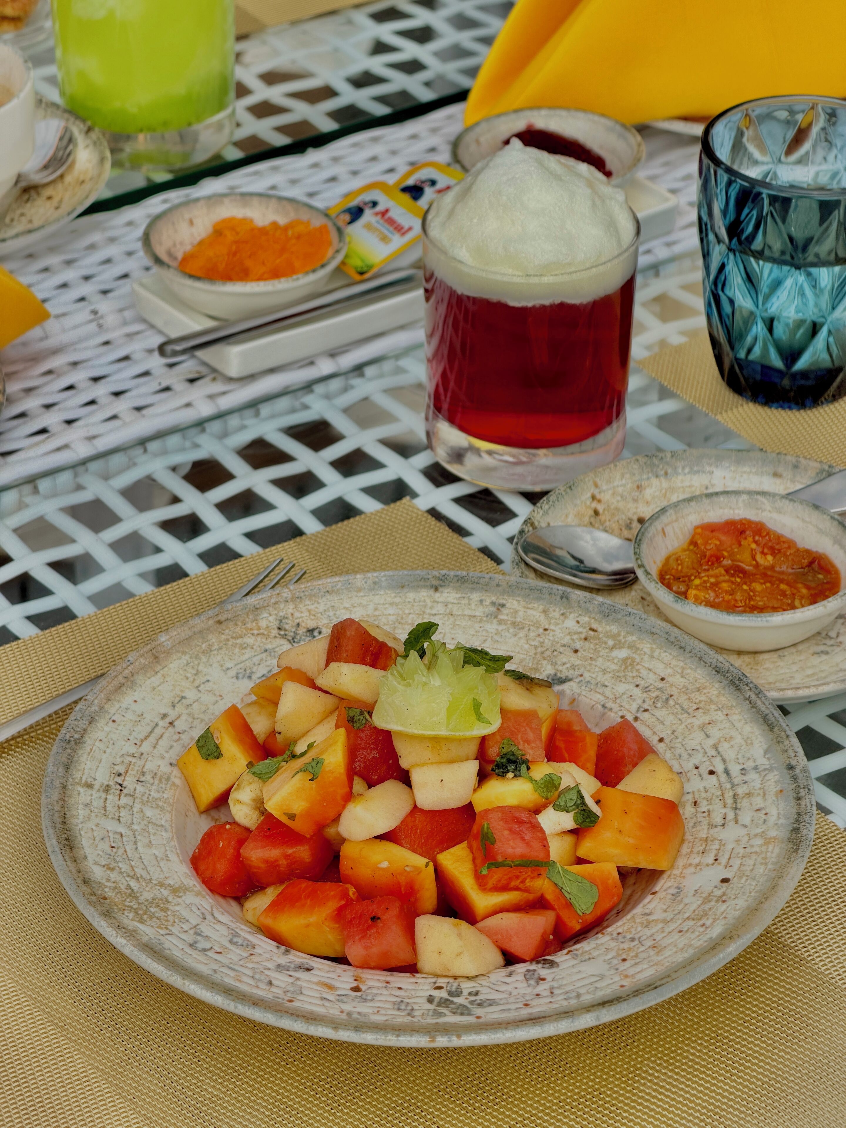 Una mesa con una ensalada de frutas, un vaso de bebida roja y diferentes condimentos.