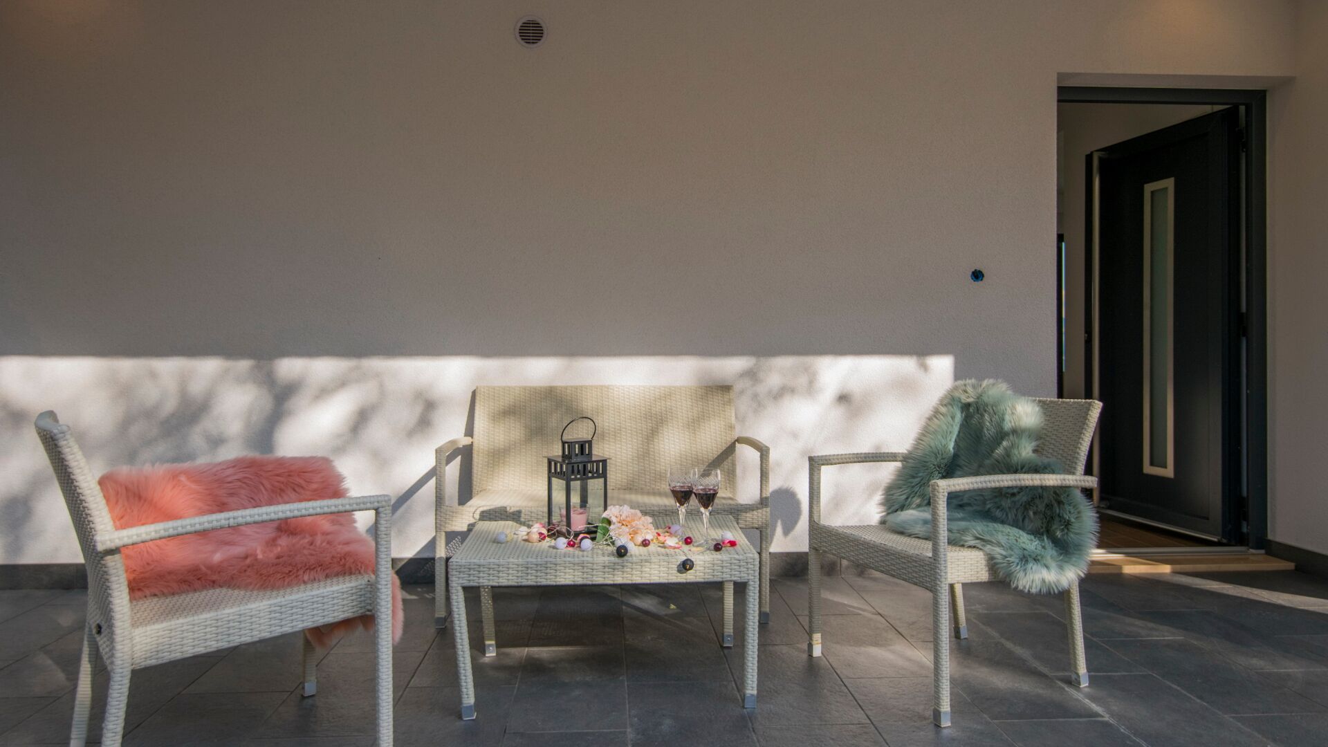 A seating area with wicker chairs and a table, a small lantern, and two glasses of red wine.