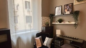 Un soggiorno accogliente con una poltrona nera, un tavolino in vetro e un piccolo divano. Si vede una finestra con vista sugli edifici, una mensola a parete con piante e un termosifone sotto la finestra.