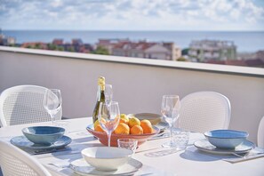 Une table dressée pour un repas avec vue sur la mer, comprenant un saladier rempli d’oranges et une bouteille de champagne.