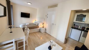 Habitación compacta con cama, mesita de noche, un pequeño escritorio con silla, una mesa blanca con un jarrón, kitchenette con armarios y microondas, y una puerta con picaporte.