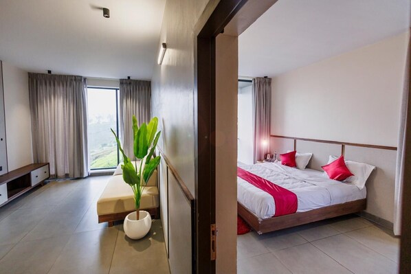 A modern hotel room with a large bed, a bedside table, a potted plant, and a view of the city through the window. - Gypsie Cottage 2 (Ootacamund)