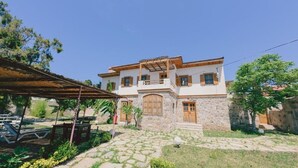 Ein zweistöckiges Steinhaus mit einem Balkon, einer Holzpergola und einer Steintreppe, die zum Eingang führt.