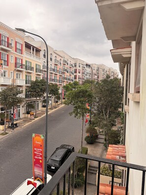 Un balcón con vista a una calle con edificios de varios pisos, un coche estacionado y una bandera.