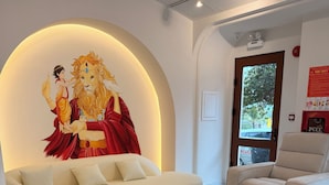Un hall d’hôtel moderne avec un canapé blanc, une table basse ronde en bois et un fauteuil beige. Une grande fresque représentant un lion et un enfant est illuminée par un luminaire circulaire.